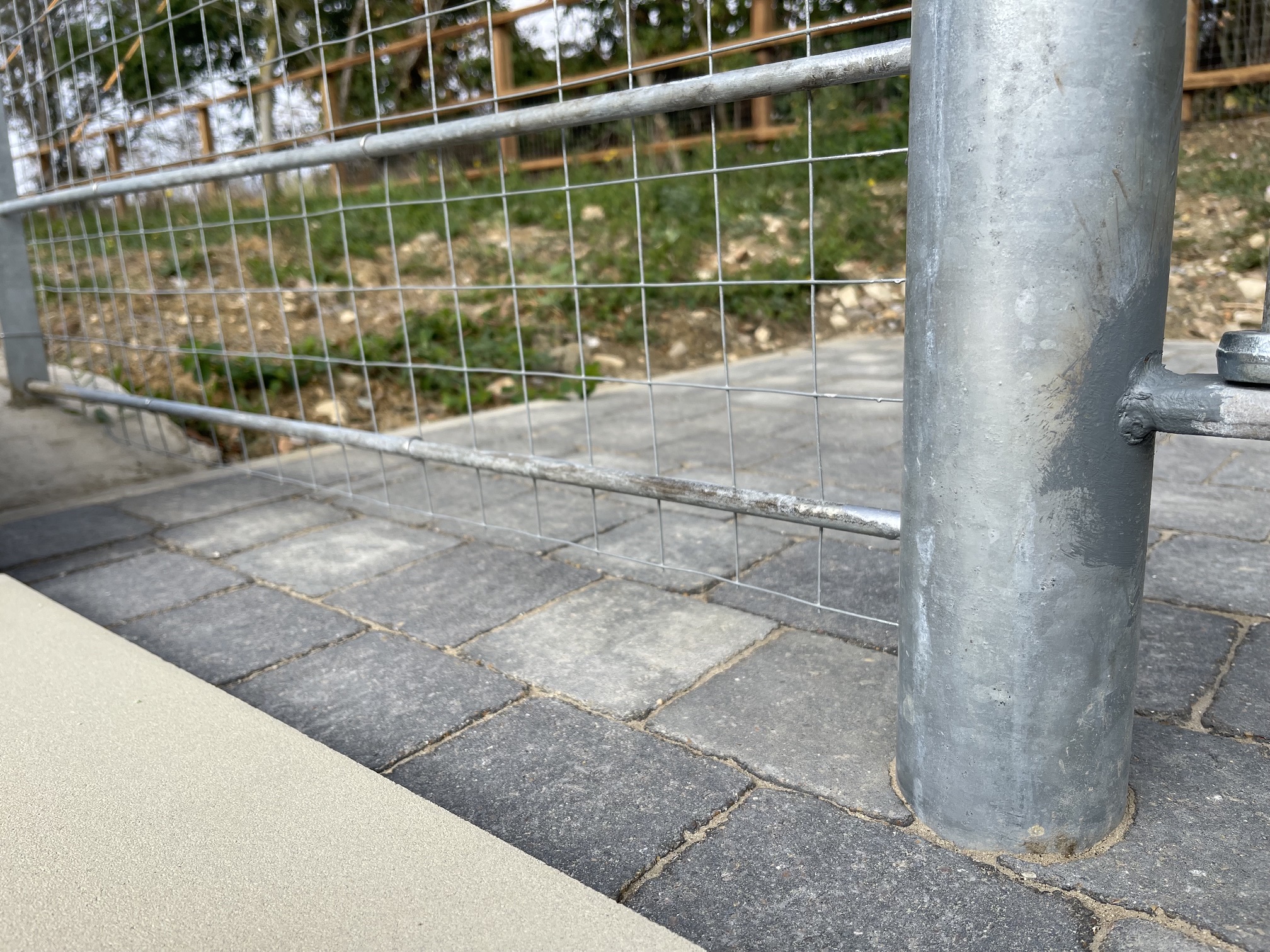 Detail of a galvanised steel railing post and wire mesh meeting block paving
